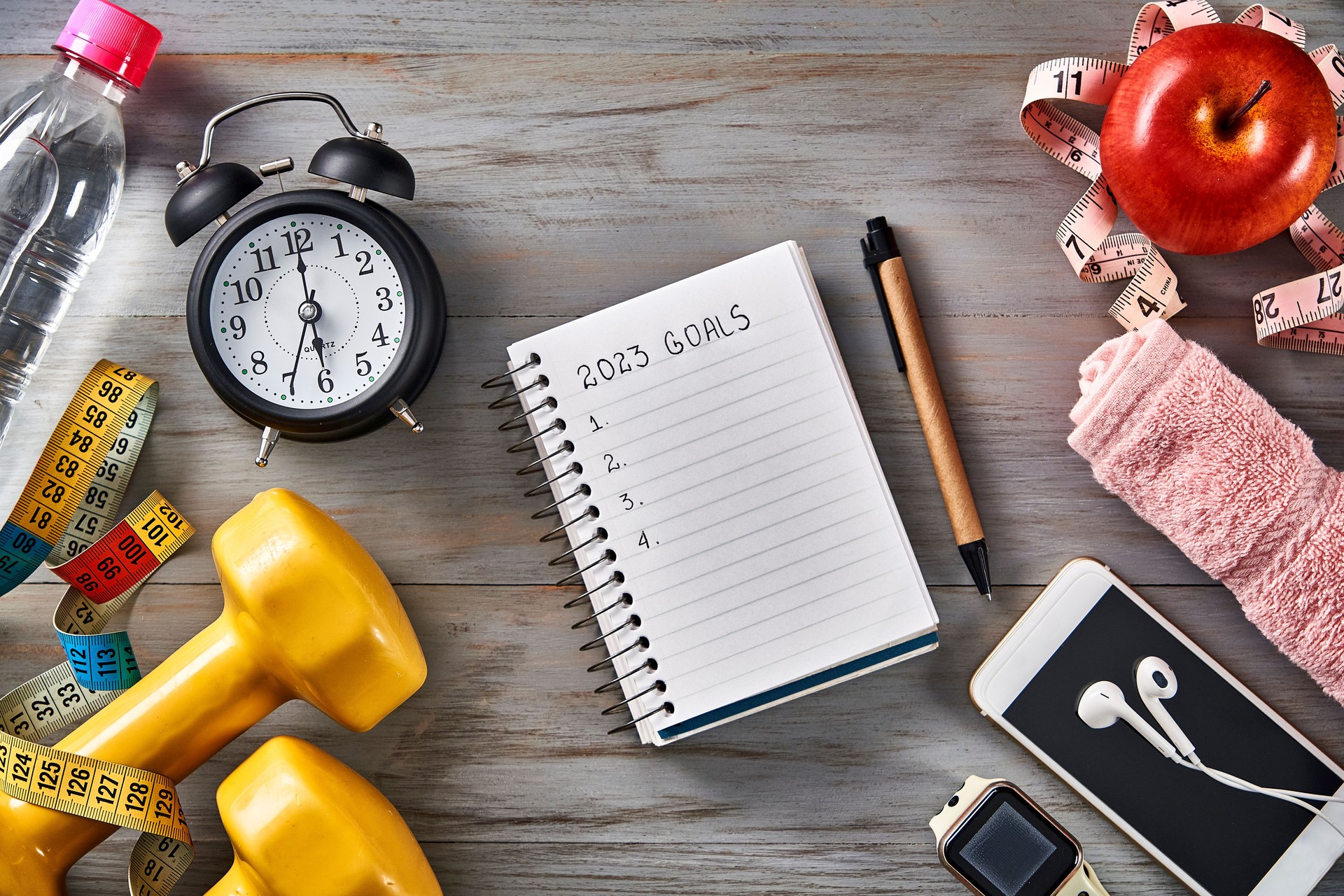 Top view of Clock, notepad with pen, tape measure, water bottle, apple, towel and dumbbells on a table